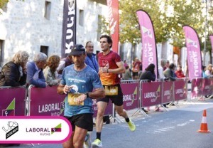 Dos corredores con dorsal y público animando tras las vallas